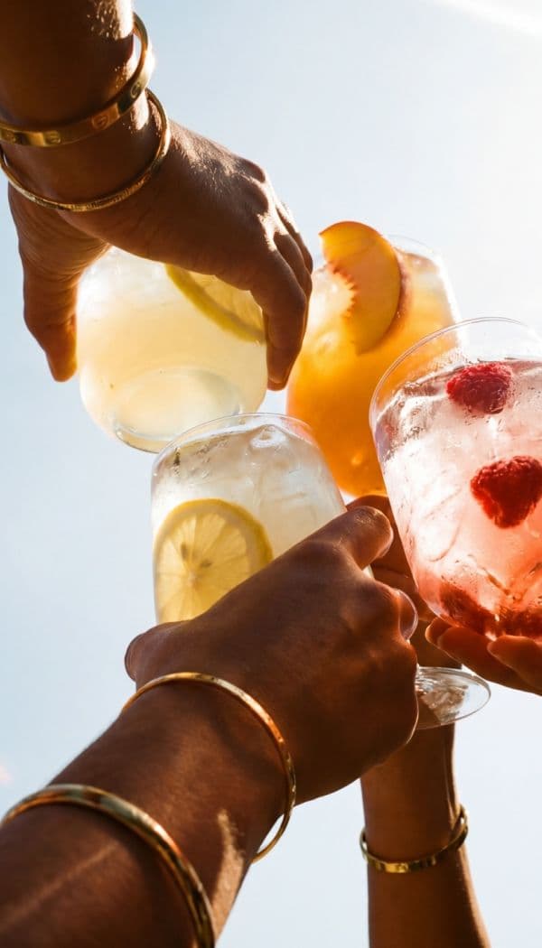 Hands in gold bangles clink fruit-garnished cocktails skyward in bright sun, a celebratory toast